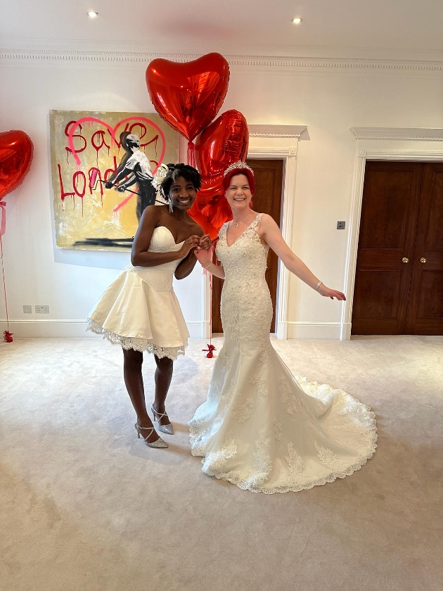 Two women wearing wedding dresses while holding red, heart-shaped balloons