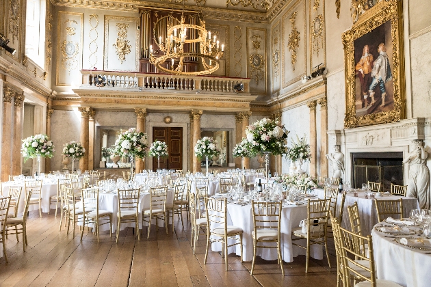 A grand room filled with gold detailing, round white tables and flowers