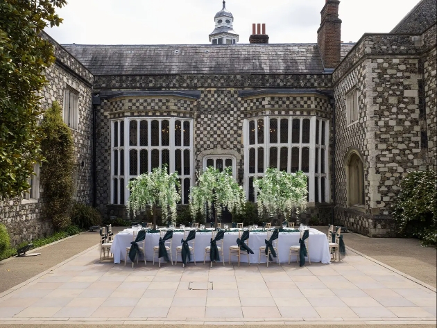 A long table white a white table cloth and flowers sitting outside in a large courtyard