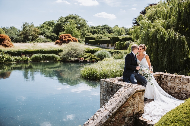 A bride and groom sitting on a wall with a pond behind them