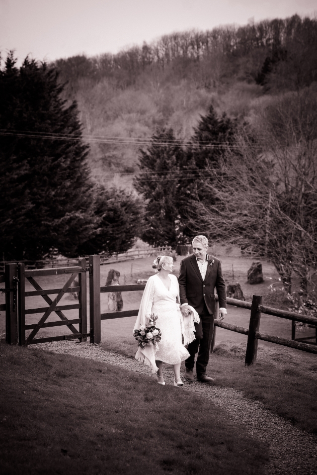 A black and white image of a bride and groom walking hand-in-hand in a field